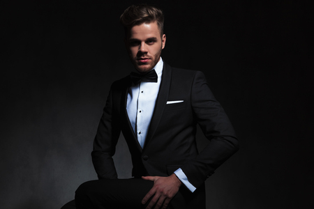 side view of young man dressed formally sitting on black background while holding his thighの写真素材