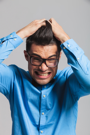 desperate young student with glasses pulling his hair with both hands while standing on light grey backgroundの写真素材