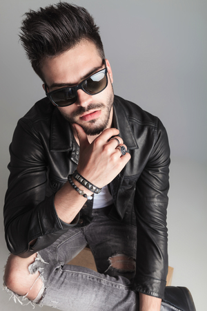 portrait of sexy man with sunglasses thinking while sitting on a wooden box on light grey background. He is wearing a leather jacket and a pair of grey ripped jeansの写真素材