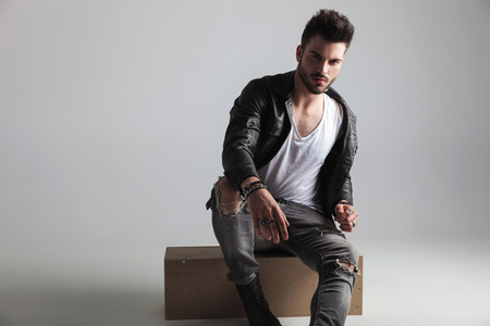 sexy man wearing leather jacket and grey jeans sitting on a wooden crate on light grey backgroundの写真素材