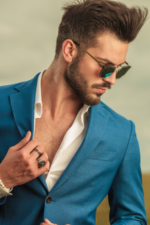 portrait of sensual man with sunglasses holding suit collar and looking down to side while standing outside on field, on cloudy weatherの写真素材