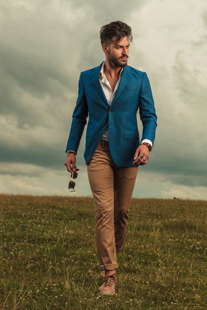 smart casual man walking and looking to side while holding sunglasses on grass field on a cloudy weather, full body pictureの写真素材