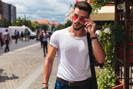 portrait of sexy man fixing sunglasses and looking to side while standing on the street, outside in the cityの写真素材