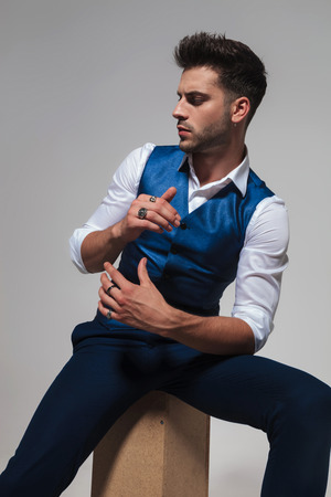 portrait of stylish man wearing a blue waistcoat sitting on a wooden box and looking down to side on light grey backgroundの写真素材