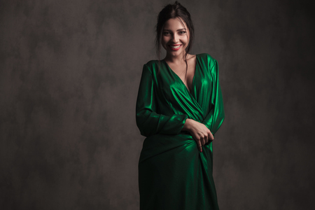 smiling brunette woman covering herself with her green dress while standing on grey wallpaper backgroundの写真素材