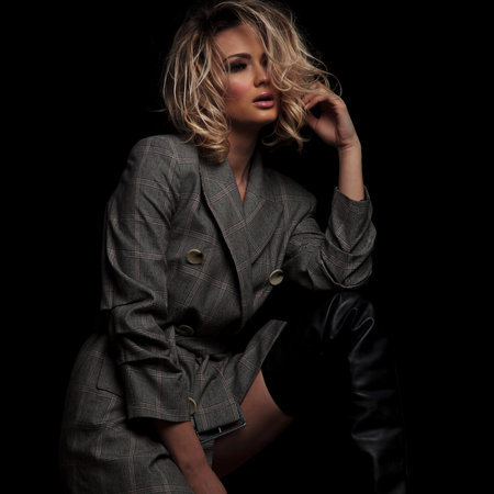 portrait of sensual blonde woman in grey suit looking to side while fixing her hair and standing...