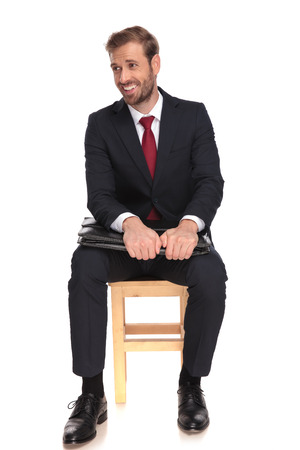 businessman sitting on wooden chair and holding briefcase under his elbows laughs and looks to side, on white backgroundの写真素材