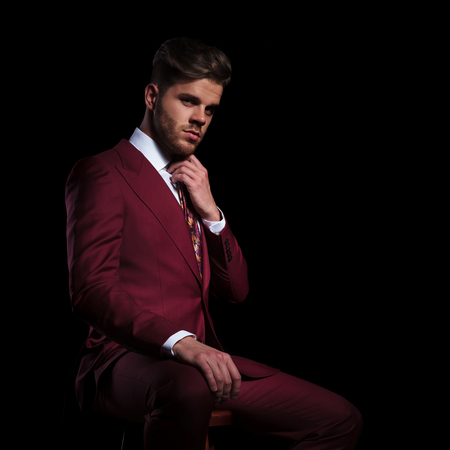seated young elegant man fixing his tie on black backgroundの写真素材