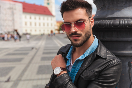 portrait of smiling casual man in leather jacket and sunglasses standing on urban background and leaning against lighting poleの写真素材