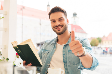 portrait of seated and relaxed casual man reading book and making ok sign in the city centerの写真素材