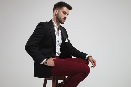 side view of seated smart casual man wearing black suit and red pants looking down on light grey background, portrait pictureの写真素材