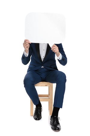 seated stylish man in blue costume covering his face with a blank talk bubble on white backgroundの写真素材