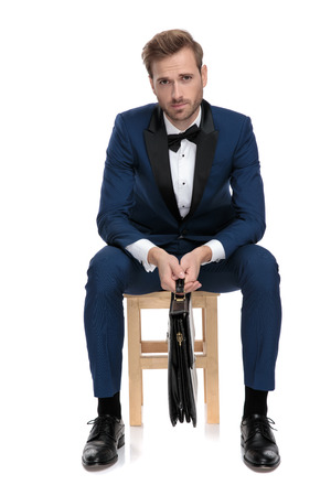 seated businessman in blue suit holds black suitcase between legs on white backgroundの写真素材