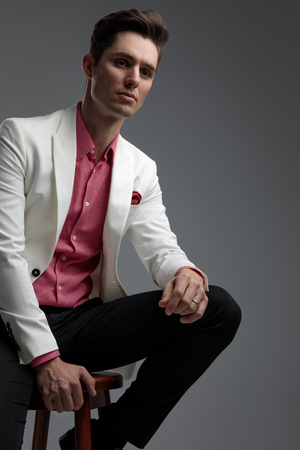 seated elegant man in suit resting his arm on leg while looking away on grey backgroundの写真素材