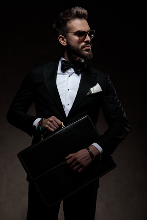 elegant man in tuxedo holds and opens suitcase while looking away to the side on dark backgroundの写真素材
