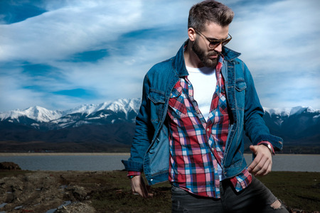 young casual bearded man moves and looks down in the outdoors, posing against lake, mountains and blue skyの写真素材