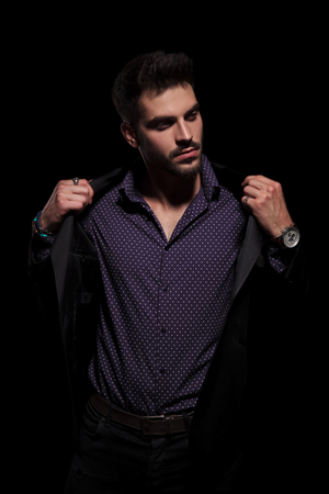 Handsome young man looking sideways and fixing his black jacket while wearing a purple shirt and standing on black studio backgroundの写真素材