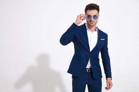Serious looking guy adjusting his sunglasses while wearing a blue suit and standing on white studio backgroundの写真素材
