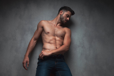 Sexy topless guy looking to the side and adjusting his jeans while leaning against gray studio backgroundの写真素材