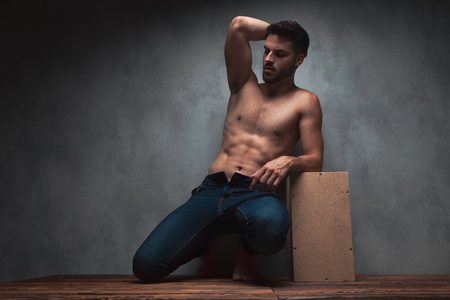 Sensual young guy kneeing and holding one of his hands on his head while wearing only jeans and leaning on a box, on gray studio backgroundの写真素材