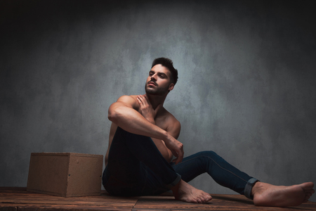 Thoughtful man looking to the side and holding one hand on his neck while being shirtless and sitting on gray studio backgroundの写真素材