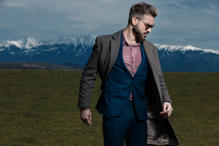 Concerned man looking to the side with his mouth open and standing while wearing a gray coat, blue suit and sunglasses on outdoor backgroundの写真素材