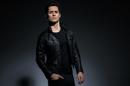Confident man posing with his hand in his pocket and looking relaxed to the camera while wearing a cap and a black leather jacket, standing on gray studio backgroundの写真素材