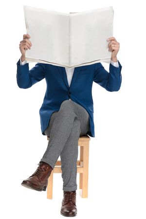 Casual man holding a newspaper over his face while wearing a suit and sitting with his legs crossed on white studio backgroundの写真素材