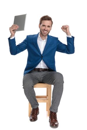 Happy casual man celebrating with his table in the air and his fist clenched while wearing a suit and sitting on white studio backgroundの写真素材