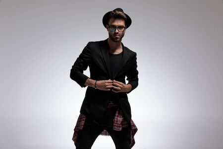 Handsome casual guy fixing his jacket while wearing a black hat and glasses, standing on gray studio backgroundの写真素材