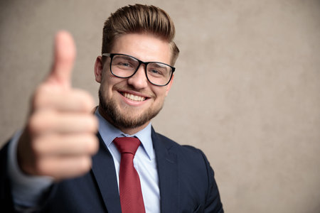 Cheerful businessman gesturing ok and smiling while wearing glasses and a blue suit, standing on...