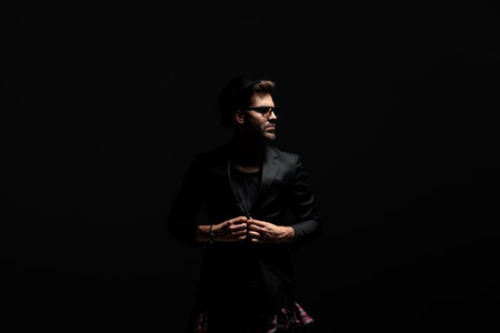 Motivated casual man unbuttoning his jacket and looking sideways while wearing sunglasses and a hat,...