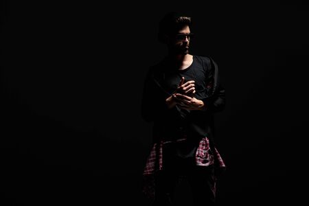 Determined dramatic guy adjusting his bracelet and looking to the side while wearing sunglasses and a hat, standing on black studio backgroundの写真素材