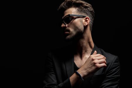 Dramatic young man looking to the side with one hand around his chest while wearing sunglasses and a black jacket, standing on black studio backgroundの写真素材