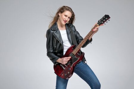Passionate young girl playing electric guitar and wearing a leather jacket and jeans while standing and posing on gray studio backgroundの写真素材