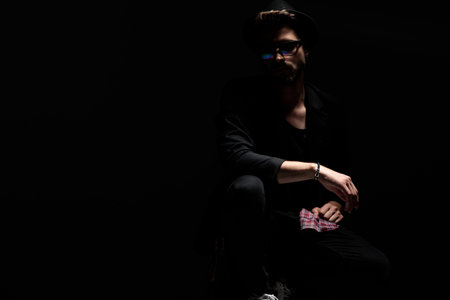 Mysterious casual man posing from the shadow while wearing sunglasses, a black hat and jacket on...