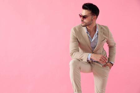 smart casual young man wearing a suit and a pair of sunglasses, leaning his elbow on his knee, standing with a hand in his pocket, looking to side on a pink backgroundの写真素材