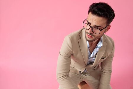 handsome man wearing glasses and suit, leaning elbows on knees, looking to side on pink background, sitting on a wooden box, portrait.の写真素材