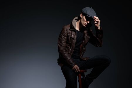side view of a handsome young man casually dressed sitting and looking ahead with shaded face while gently fixing his hat on gray studio backgroundの写真素材