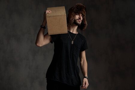attractive casual man holding a wooden box on his shoulders, wearing a fur hat, looking to side, standing isolated on grey backgroundの写真素材