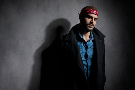 side view of attractive young man holding hands in pockets, wearing a longcoat and a red bandana, standing on grey background in studio, portraitの写真素材
