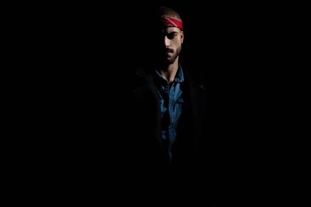 attractive young man wearing a red bandana and a long coat, half standing in light on black background, on studio, portraitの写真素材