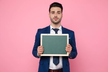 attractive young man wearing navy blue suit, holding an empty board, standing on pink background in studioの写真素材