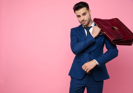 attractive young man wearing navy blue suit, holding suitcase and standing on pink background in studioの写真素材