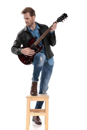 sexy casual man with black leather jacket is standing with one leg on chair and the other down while playing the guitar passionate and looking to a side on white studio backgroundの写真素材