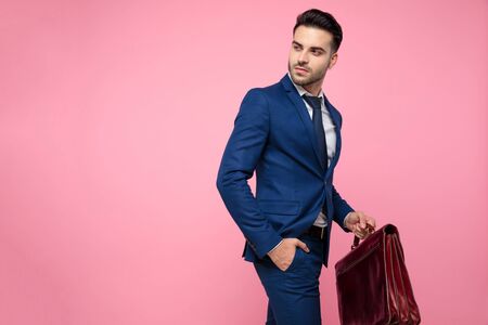 side view of attractive young man wearing navy blue suit, holding suitcase and walking with hand in pocket, looking to side and standing on pink background in studioの写真素材