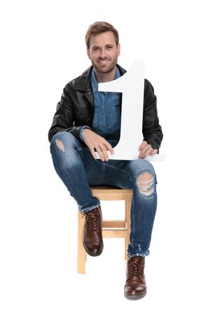 young casual man with black leather jacket is sitting on a wooden chair with a number one in hands happy on white studio backgroundの写真素材