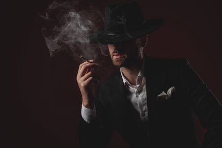 attractive dramatic man wearing tuxedo and black hat, smoking and sitting on black background in studioの写真素材