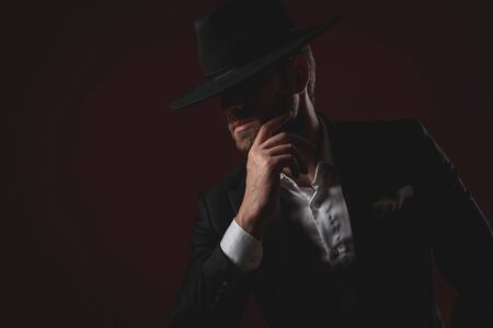 attractive young man wearing tuxedo and black hat, looking to side and holding hands to chin, sitting on black background in studioの写真素材