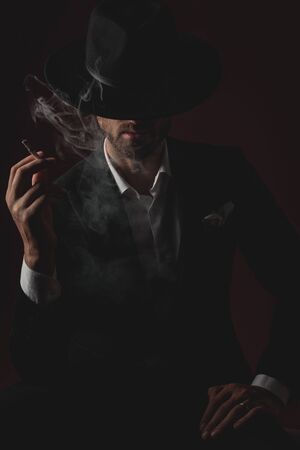 attractive young man wearing tuxedo and black hat, holding hand on knee, smoking and sitting on black backgroundの写真素材